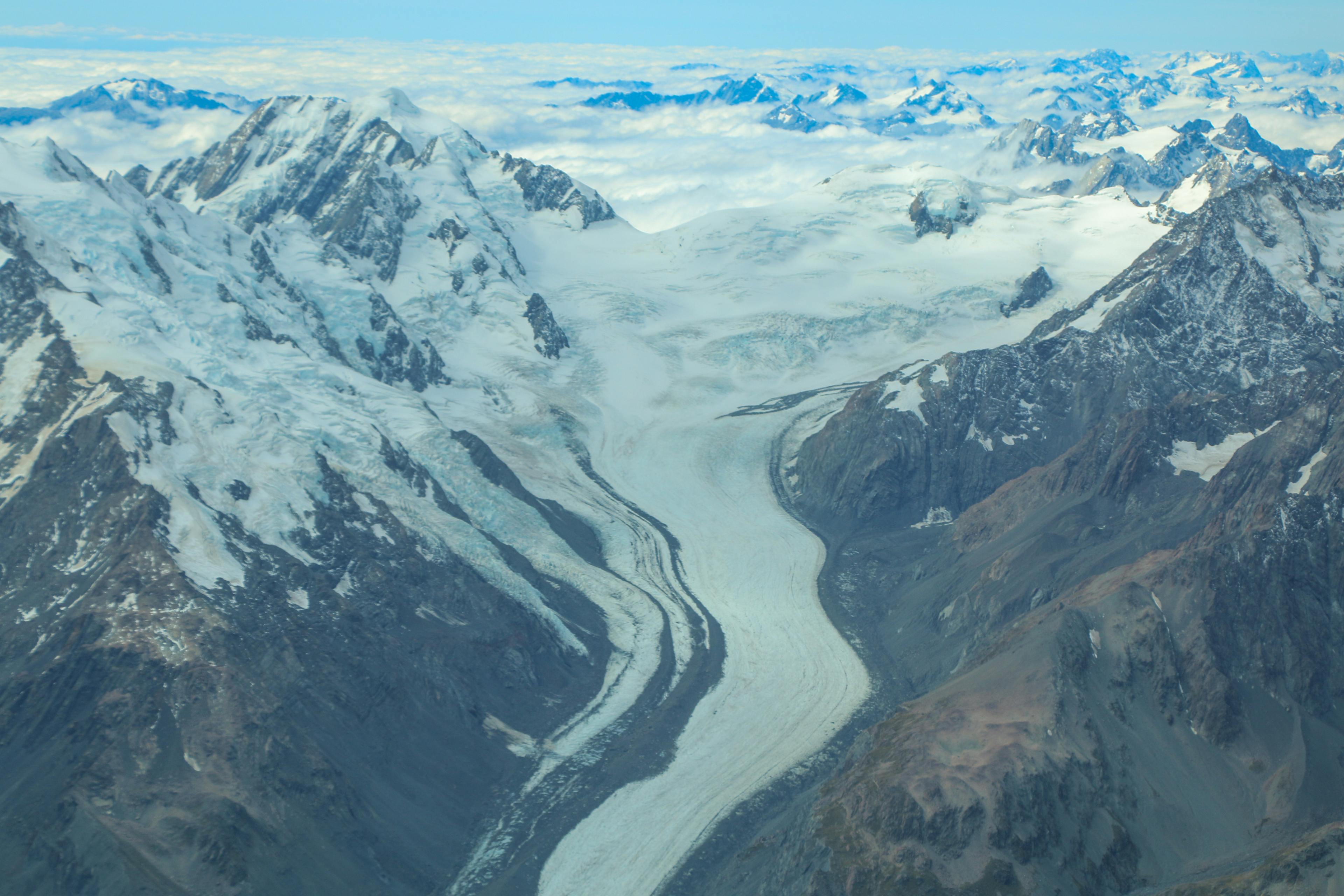 An aerial photo of a glacier snaking between tall snow-capped mountains, strewn with patches of low cloud beneath a blue sky.