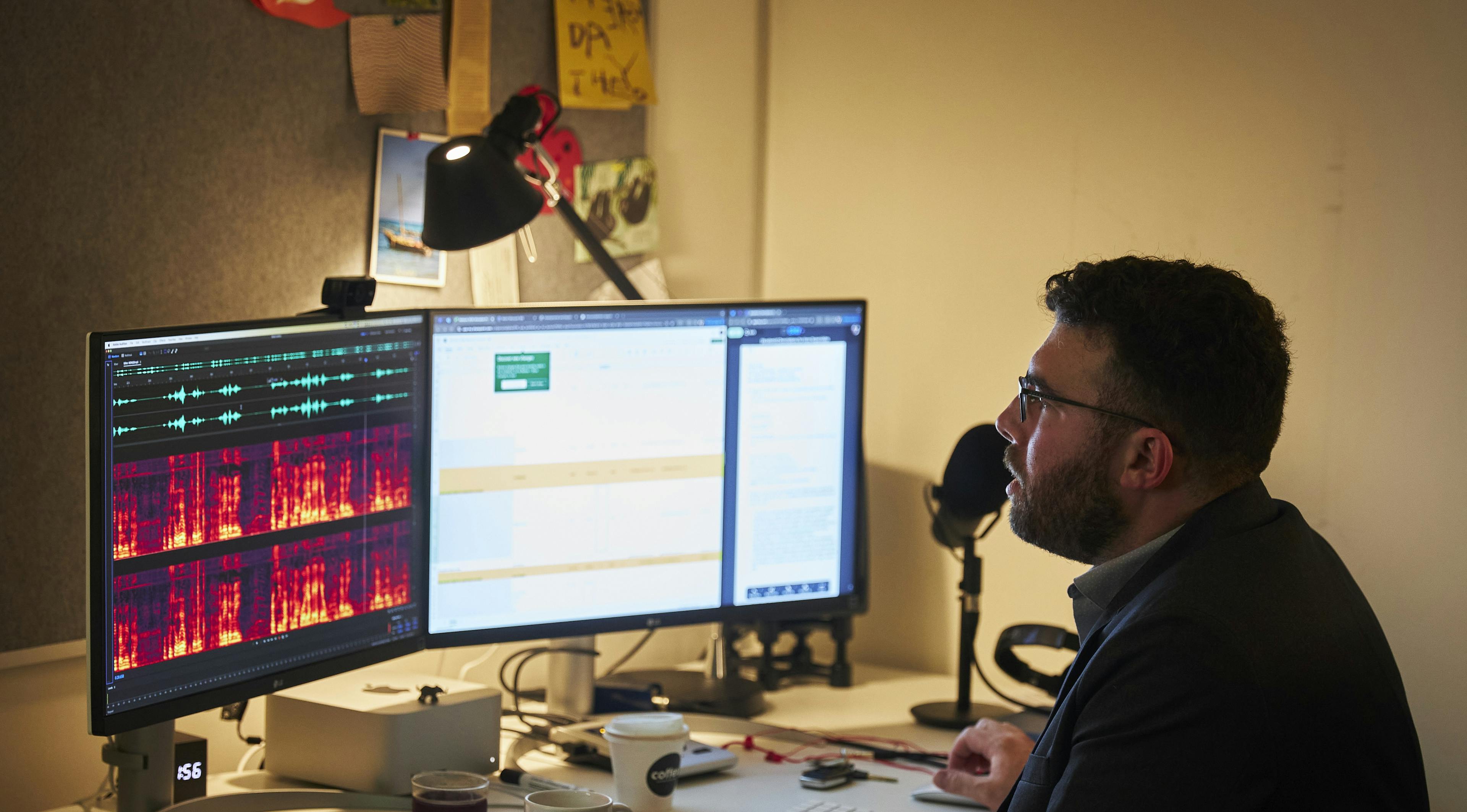 A man sitting at a desk looking at two computer monitors. One is showing brightly coloured waveforms. There is a microphone on the desk.