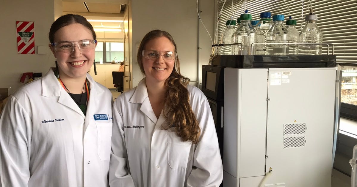 Two women in white lab coats stands in a lab next to a large machine with about a dozen large glass bottles half-filled with clear liquid sitting on top of it.