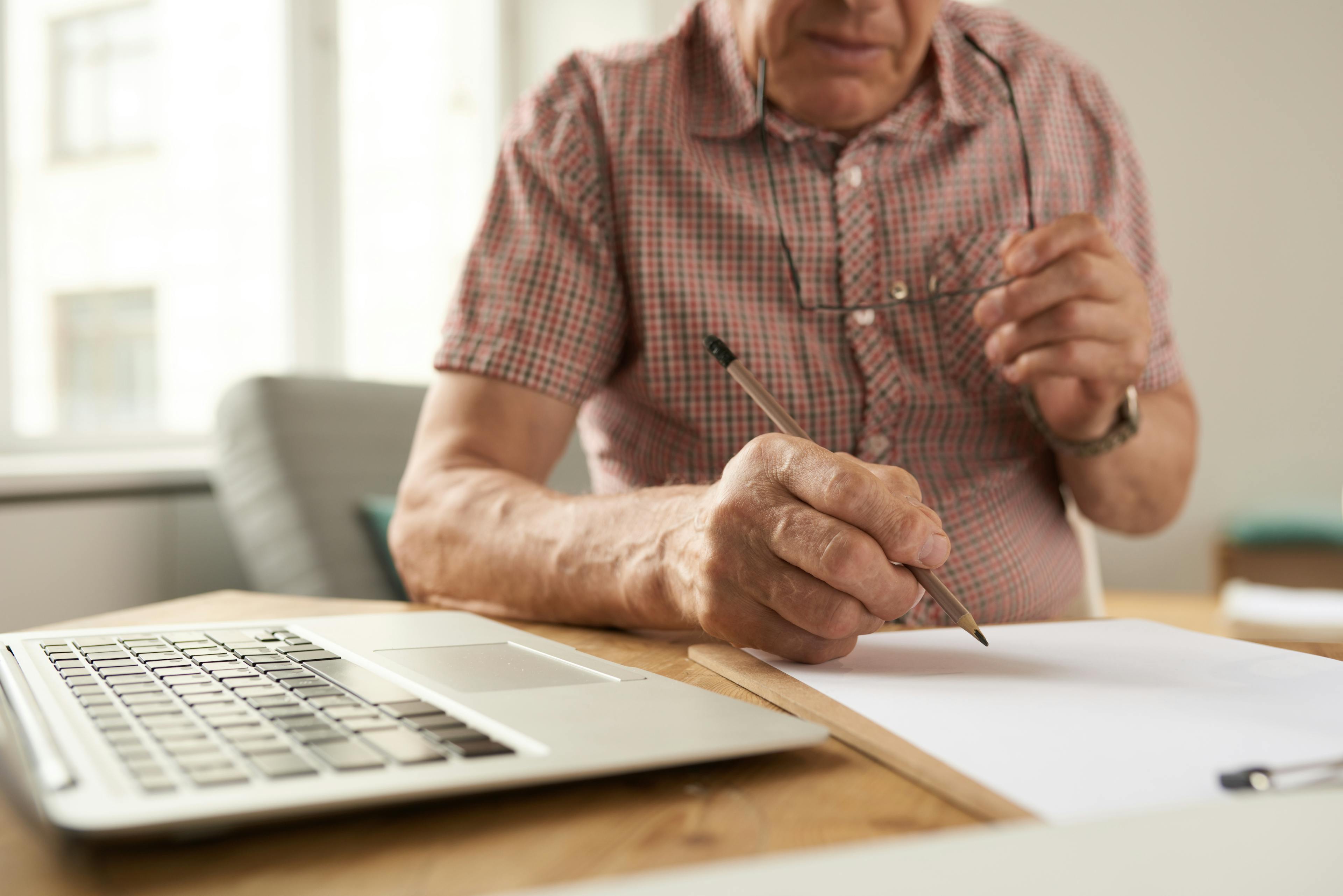 Closeup portrait of unrecognizable senior man filling in papers while working with laptop at home