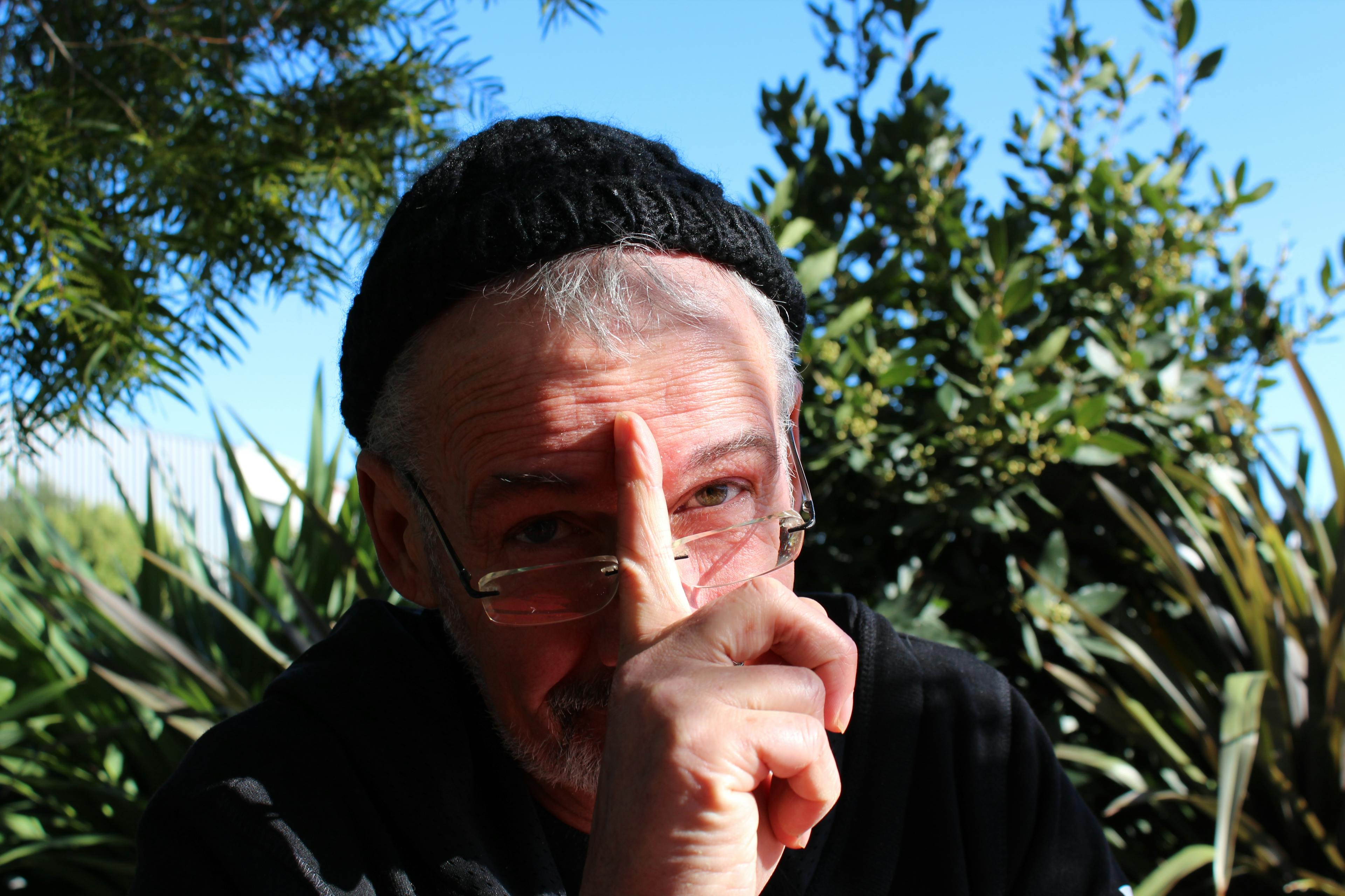 Headshot of Peter Ellis wearing a beanie with his finger held over his face, pointing skywards.