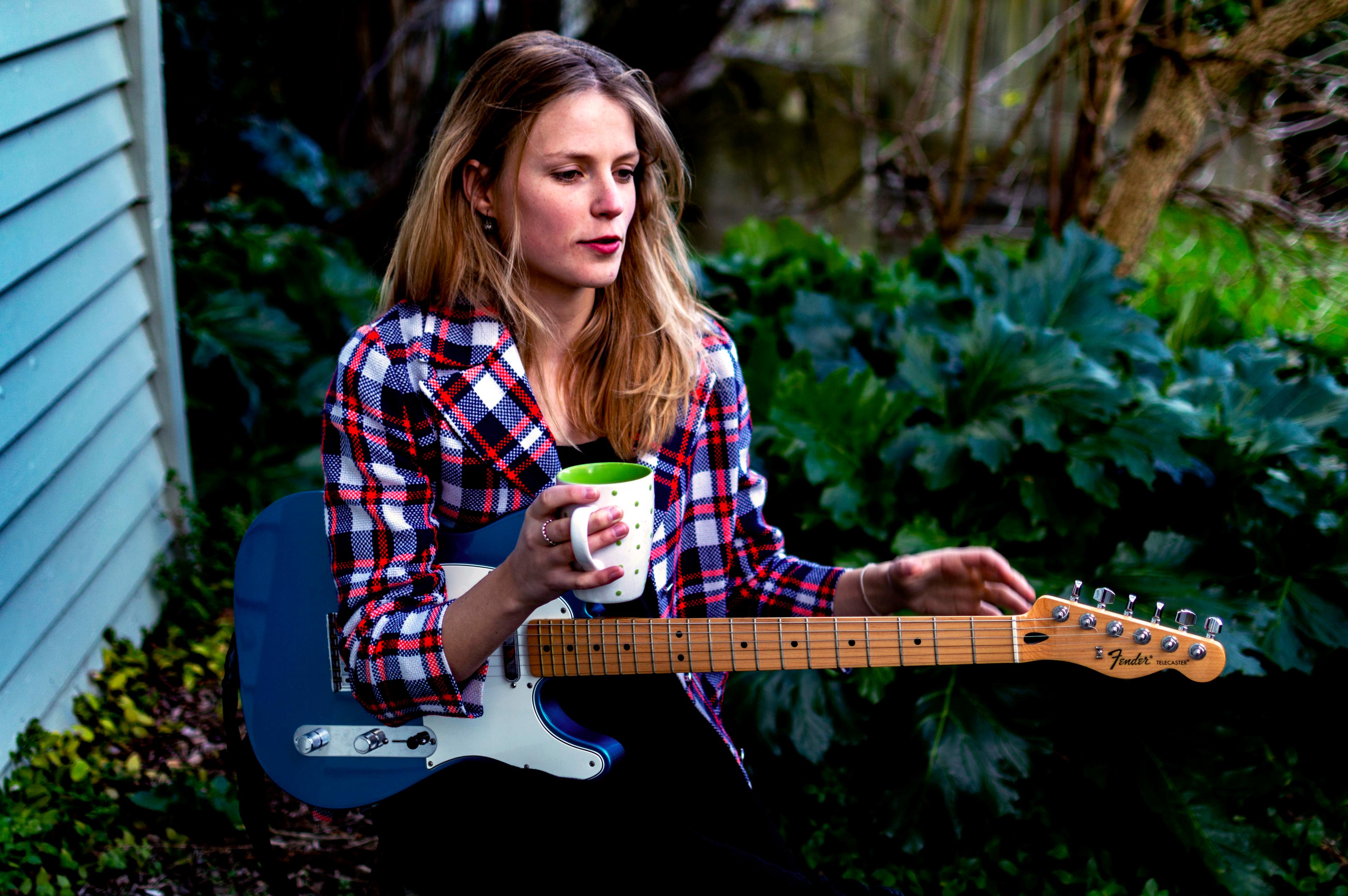 A colour photo of a young Women sitting down holding a guitar and a coffee cup