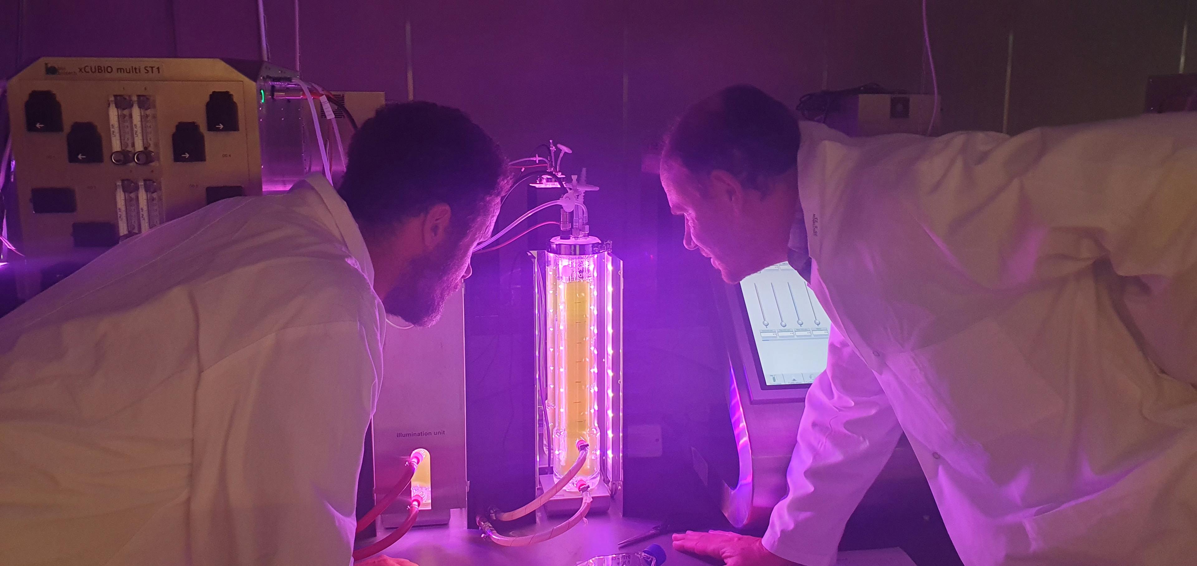 Two men in white lab coats look at a glass tube filled with something yellow-green. The room is bathed in purple light and other scientific machines are displays are visible in the background.