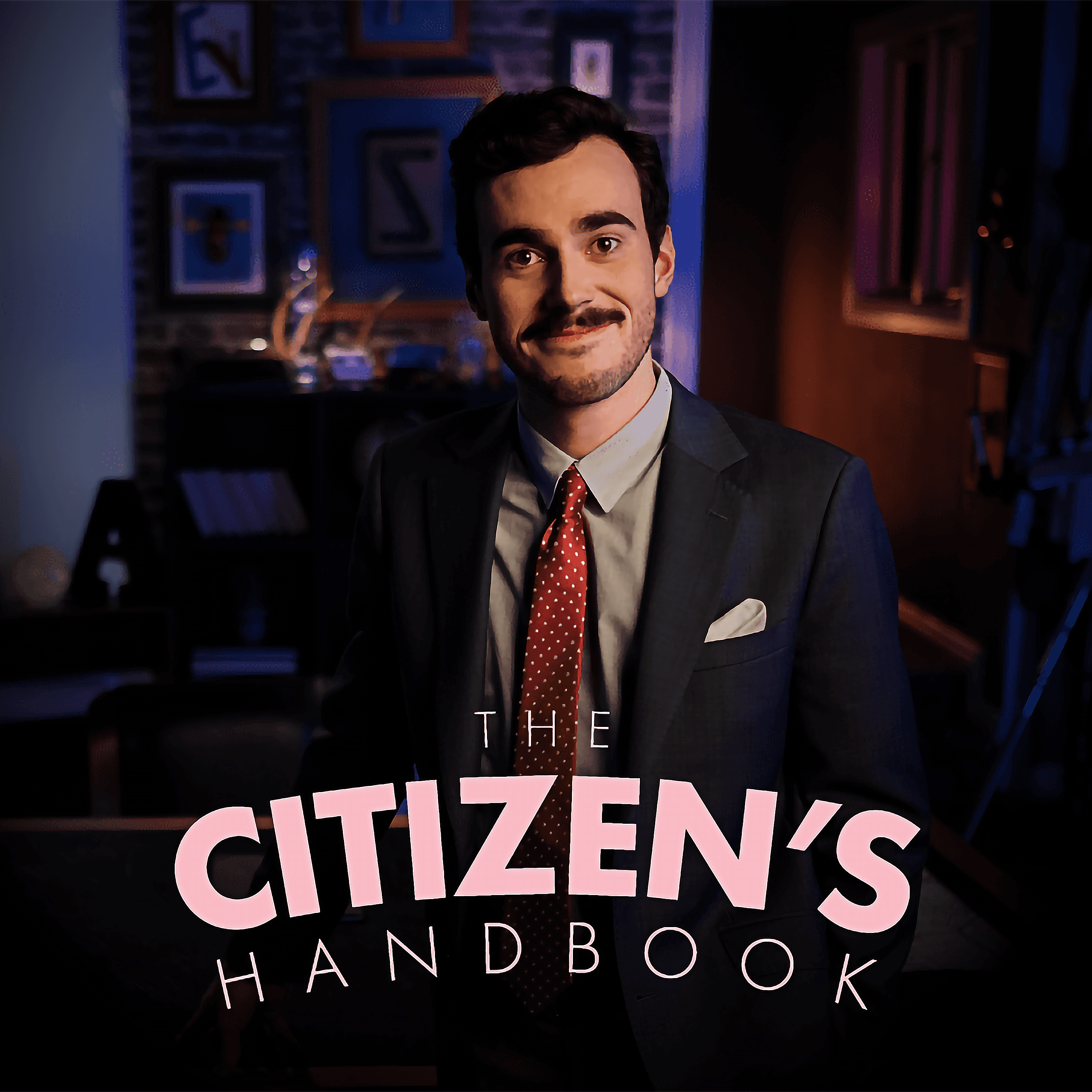 A smiling young man dressed in a suit stands before a darkly lit room with the text 'The Citizen's Handbook' appearing at the bottom of the frame.