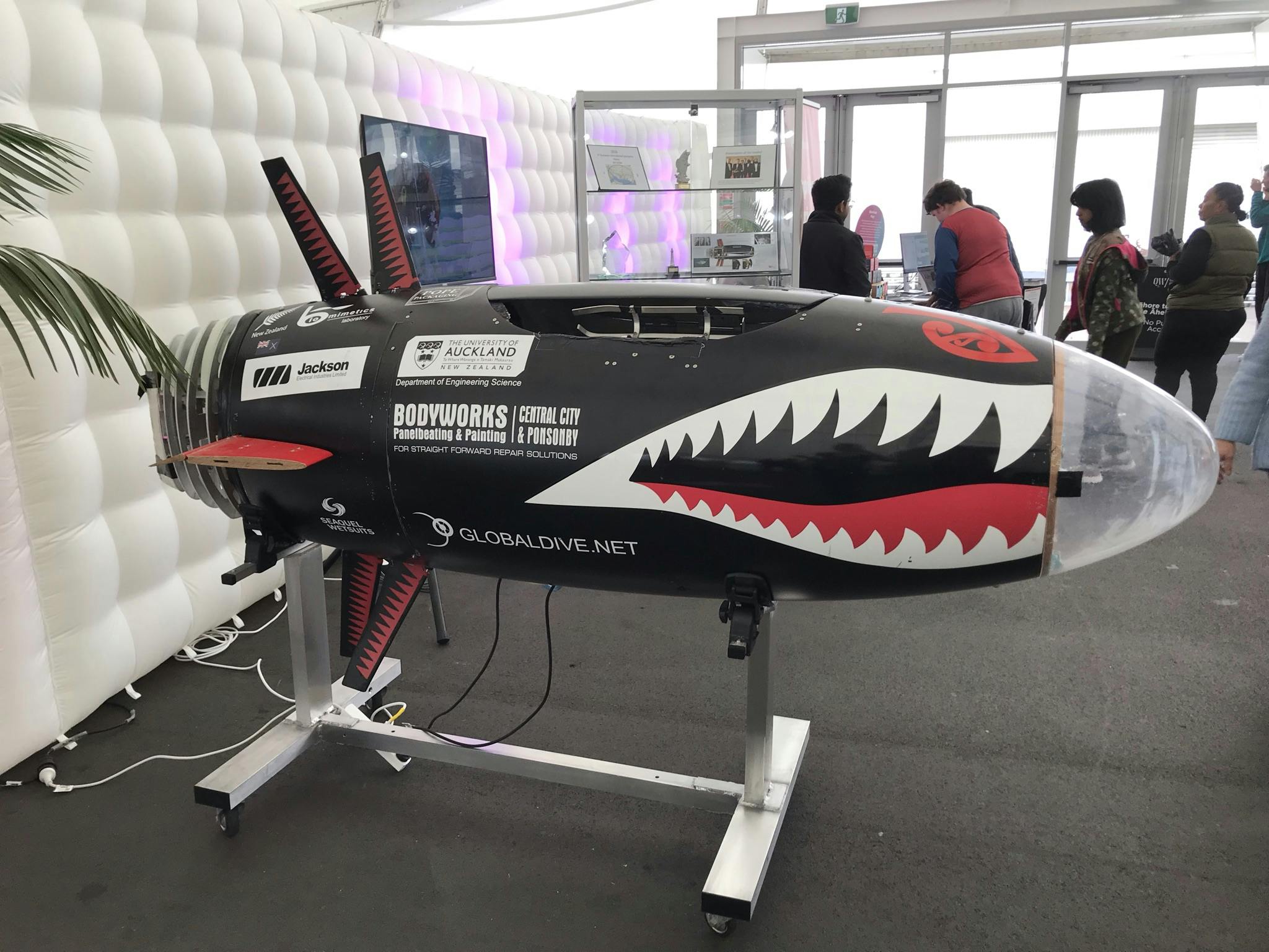 A torpedo shaped black machine sits on a silver metal stand, with sharp white teeth drawn on, and fin like things sticking out from the top and bottom.