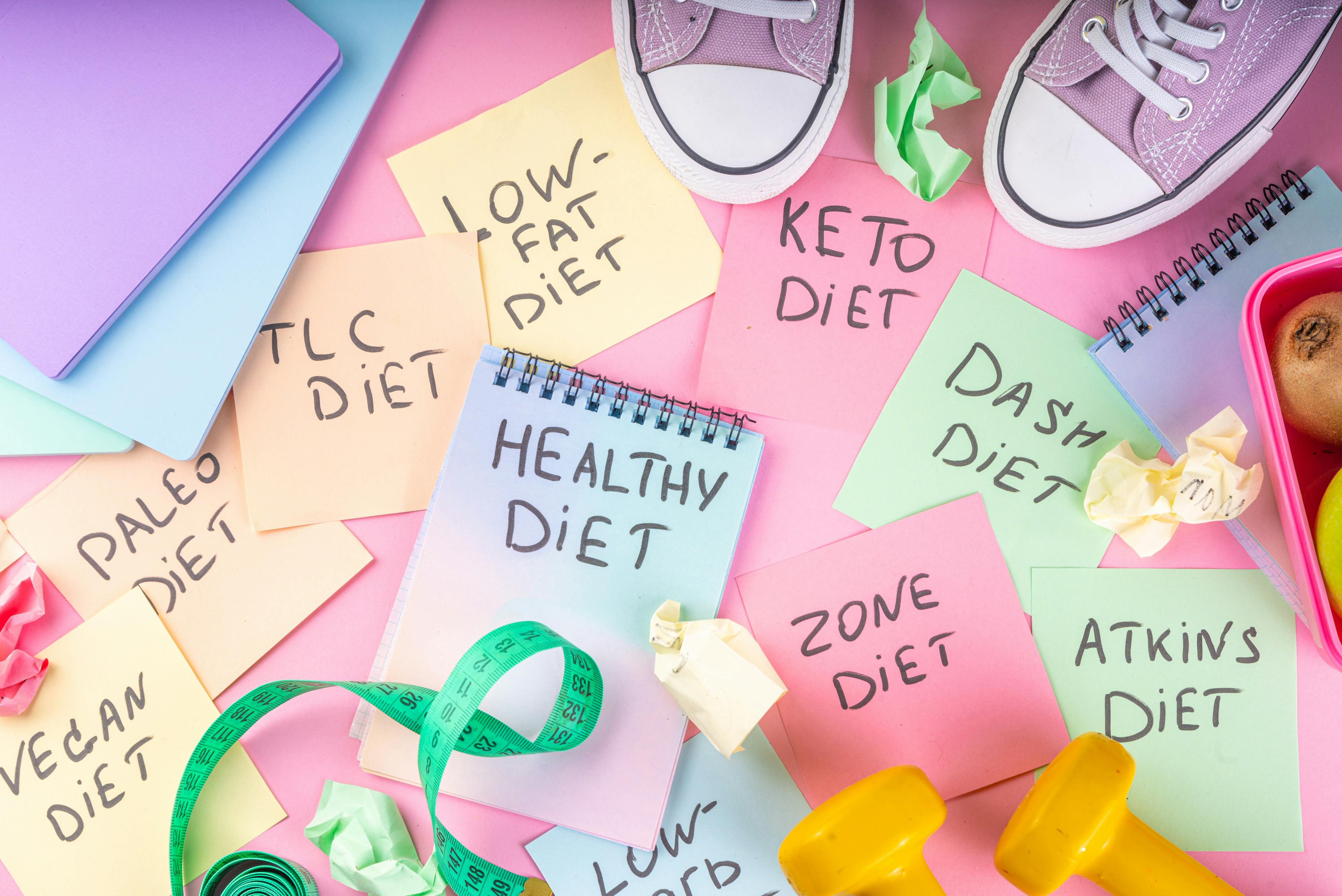 Diet choice concept with bright handwritten notes with names of trendy diets and healthy eating concept, with sneakers, tape measure, dumbbells on bright pink background.