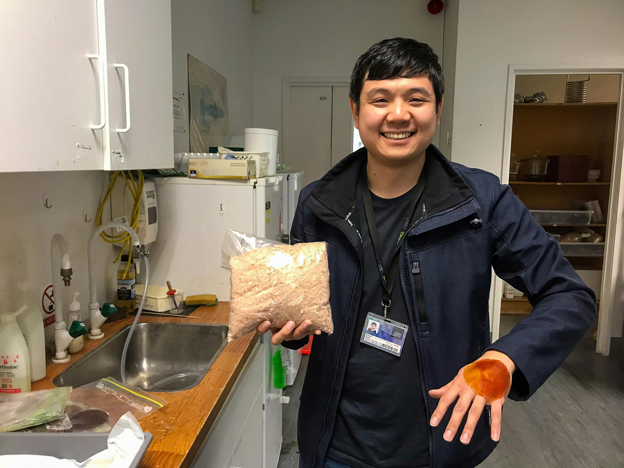 A man standing next to a kitchen sink with a fridge in the background. The man is smiling and holds a plastic bag of a coarse off-white substance and has a circular piece of orange "skin" on his outstretched hand.