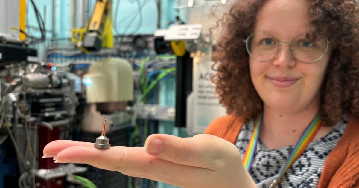 A woman with curly hair and glasses holds up a thimble-sized metal pin. She is standing in front of complicated looking machinery in a lab.