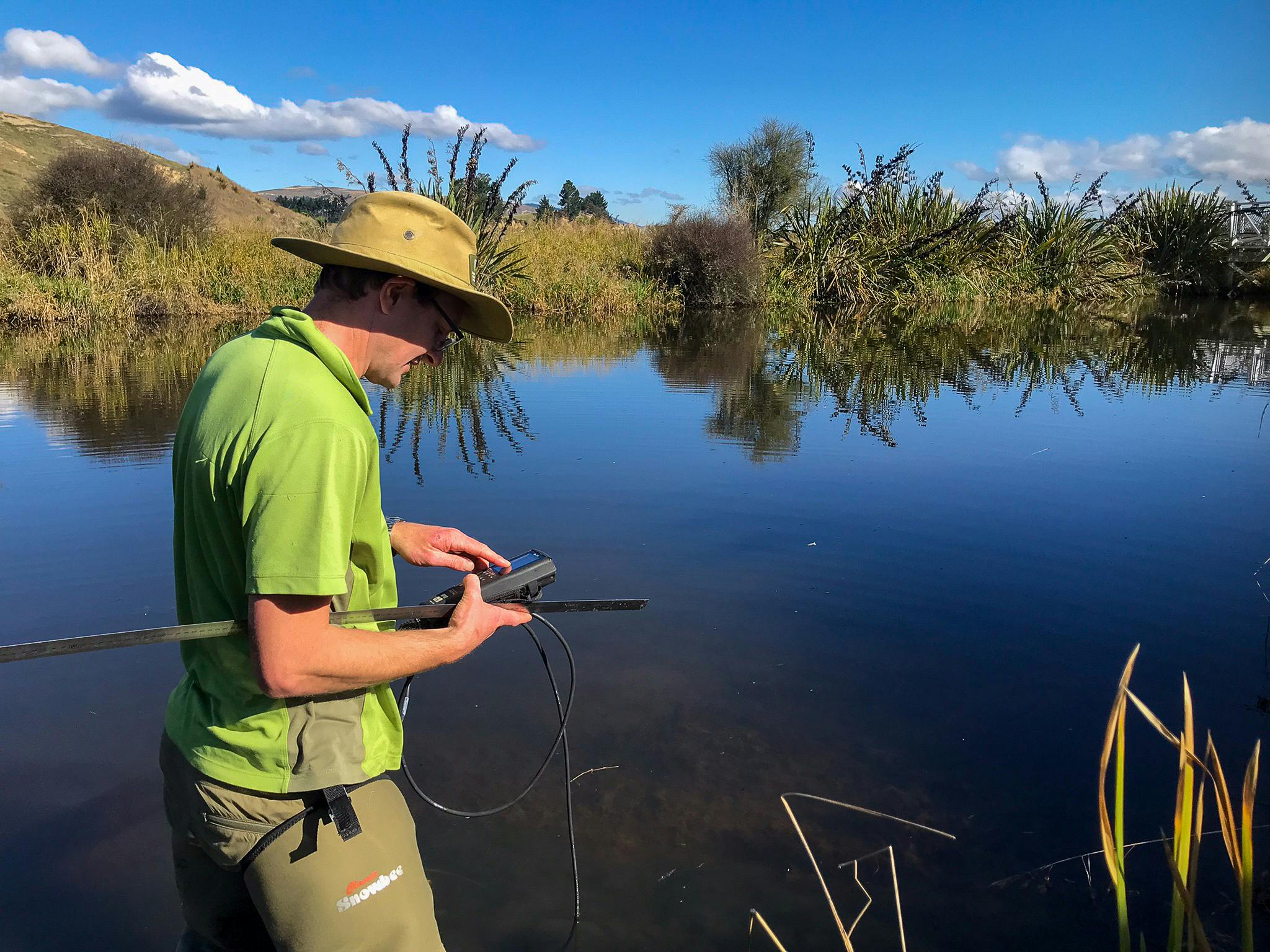 A man wearing a wide-brimmed hat and a bright green colllared t-shirt stands on the edge of a wetland pool framed by long grass and vegetation. He is side on, holding a long pole and using an electronic device with a wire extending down towards the water. It's a sunny day and the water is still.