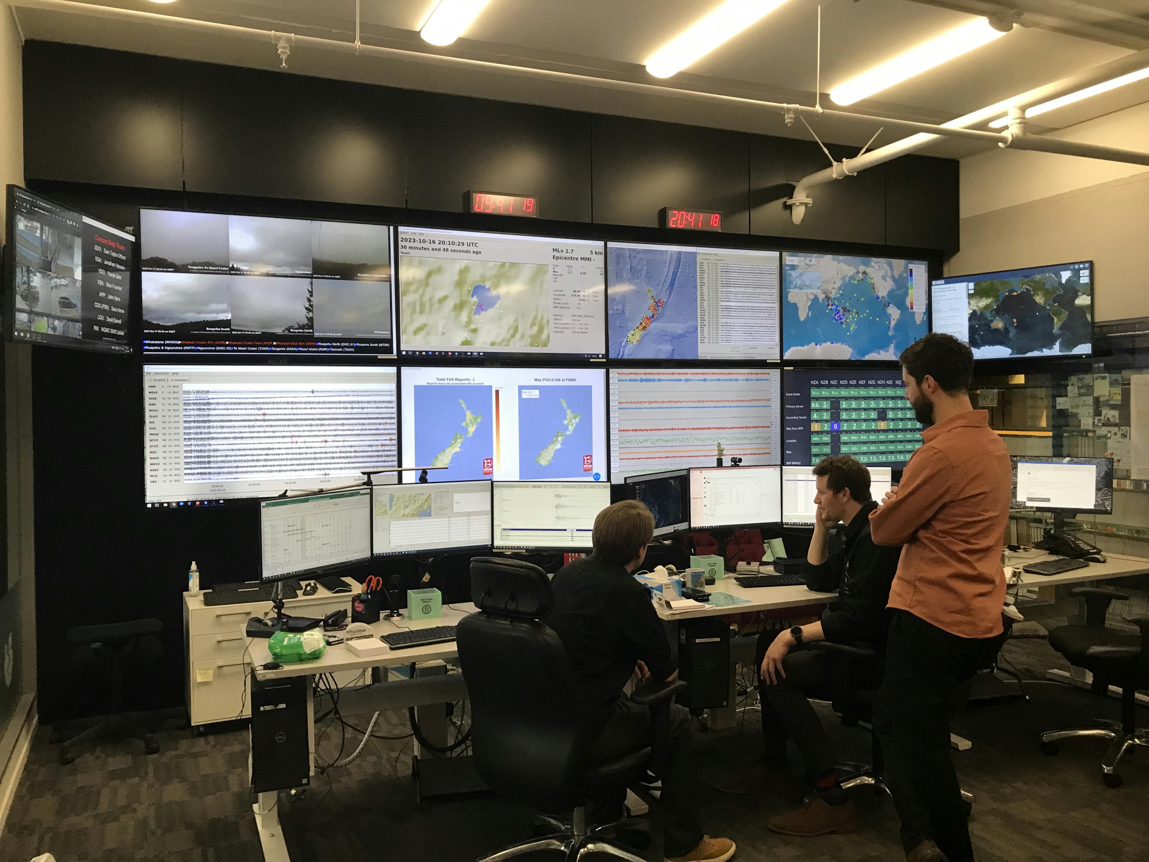 Three people look at an array of six computer monitors and 10 larger screens displaying maps and data.