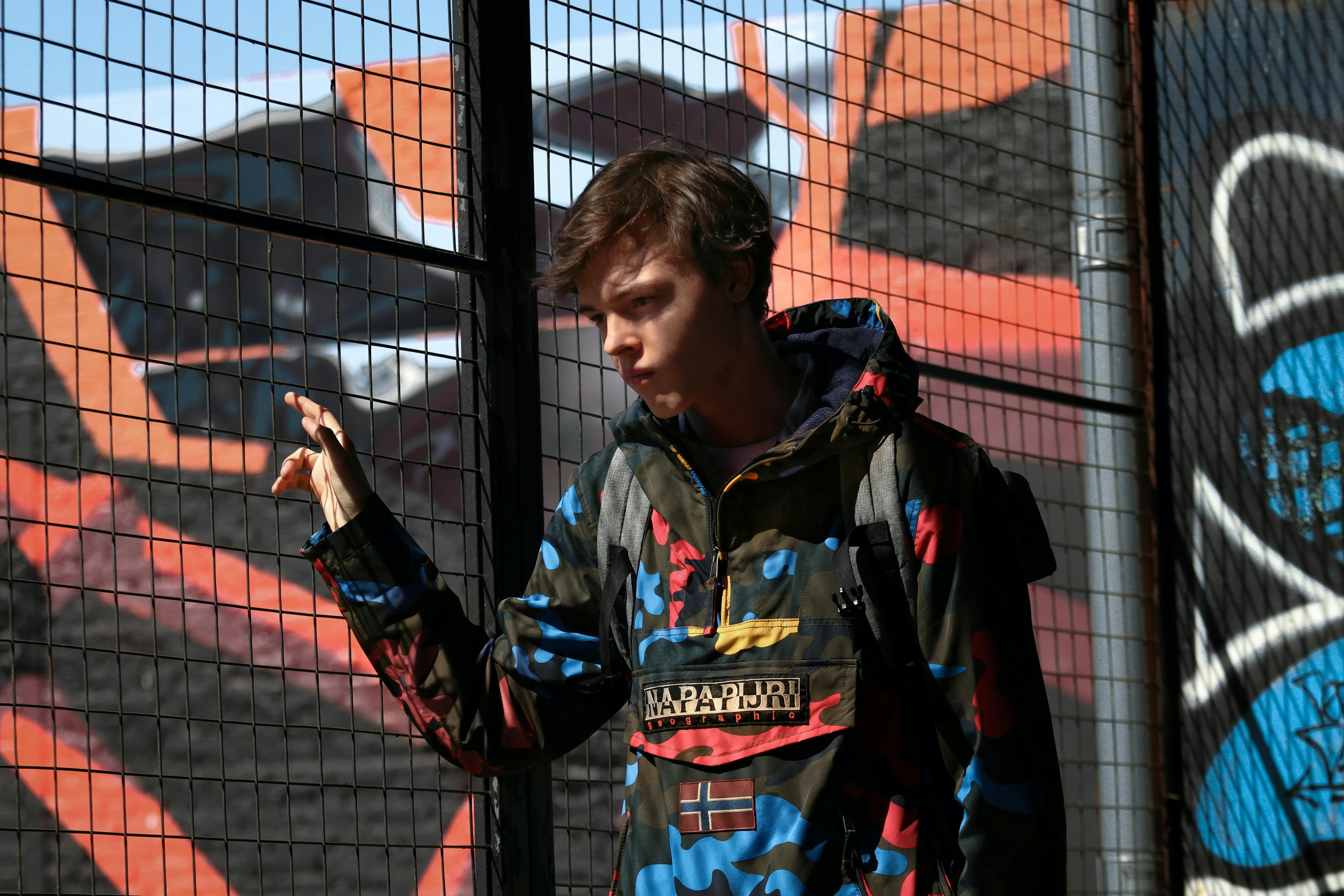 Teenage boy wearing a hoodie and backpack walks past a graffiti'd wall and wire fence