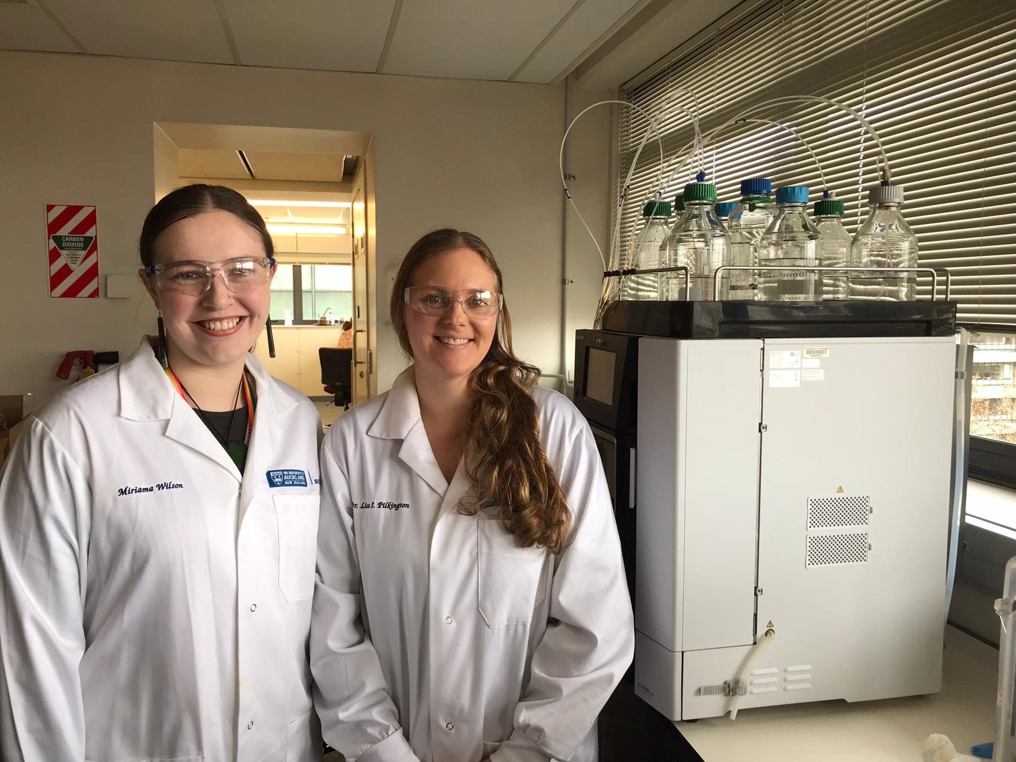 Two women in white lab coats and lab goggles stand in a lab next to a large machine with about a dozen glass bottles on top of it. The glass bottles are half-filled with clear liquid and have tubes running from them into the machine.