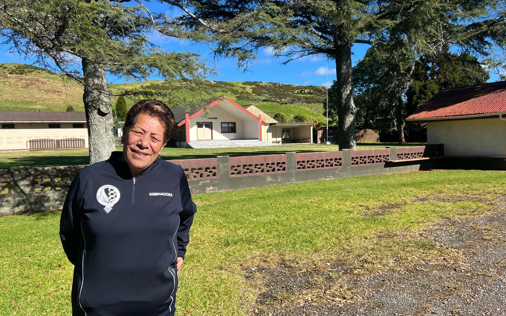 A woman in a navy blue sweater smiles while standing in front of a marae wharenui framed by two tall trees. It's a sunny day and there are green hills behind the marae buildings.