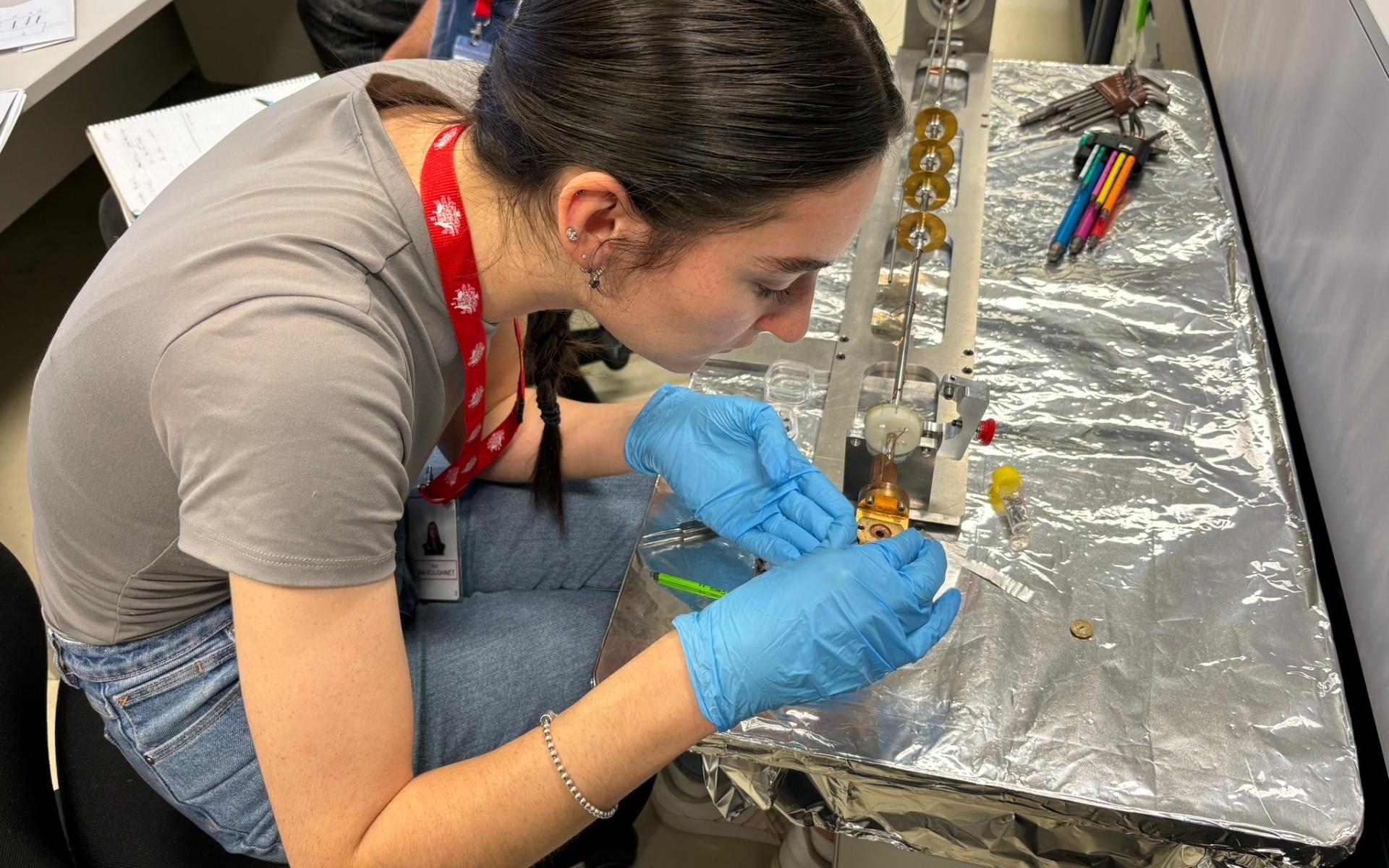 A woman wearing blue rubber gloves carefully handles a small piece of metal equipment attached to a long contraption on a bench covered with tinfoil.