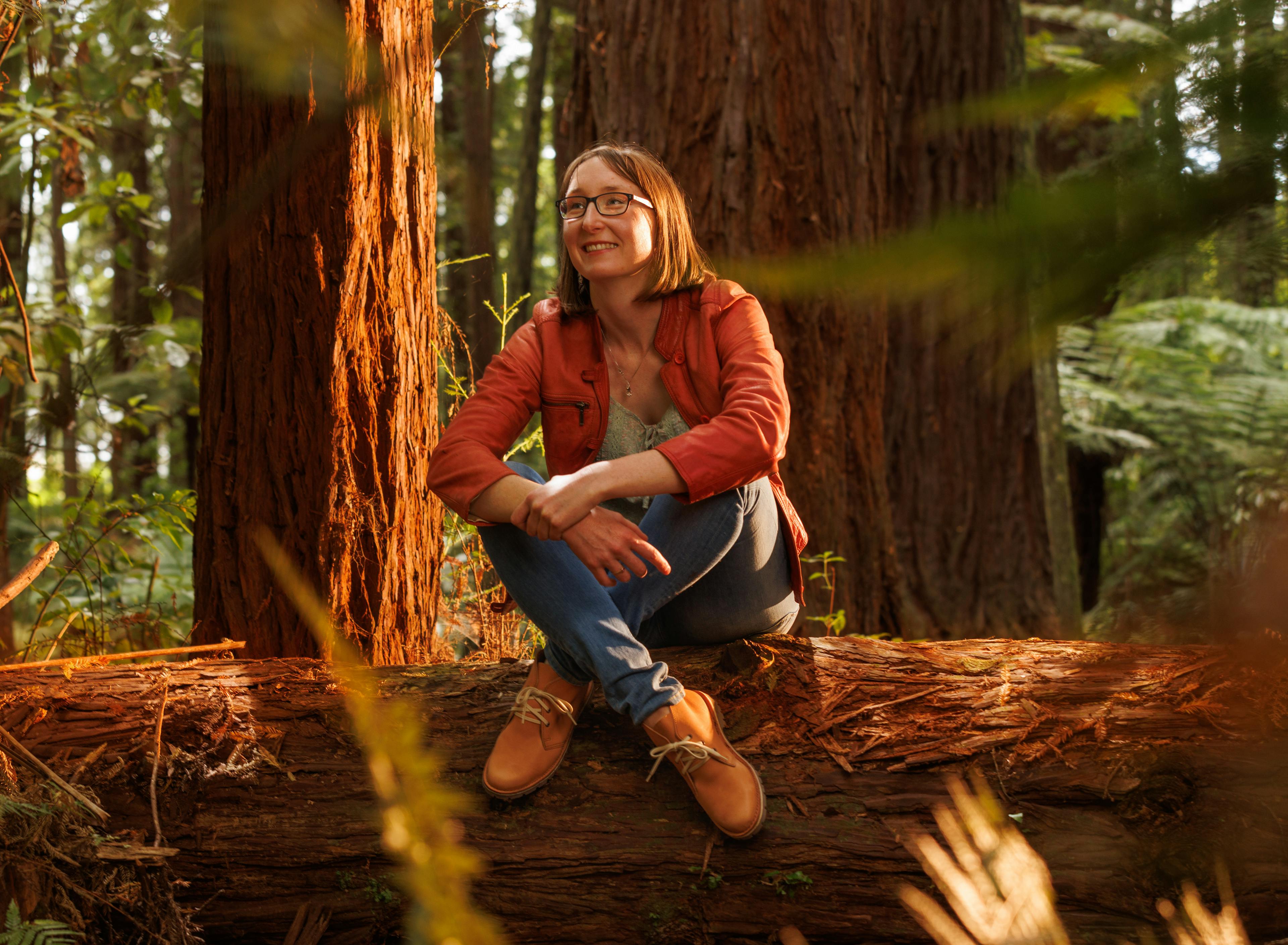 Hilary Corkran is sitting cross-legged on a fallen pine log in a forest. She is smiling and wearing tan leather boots.