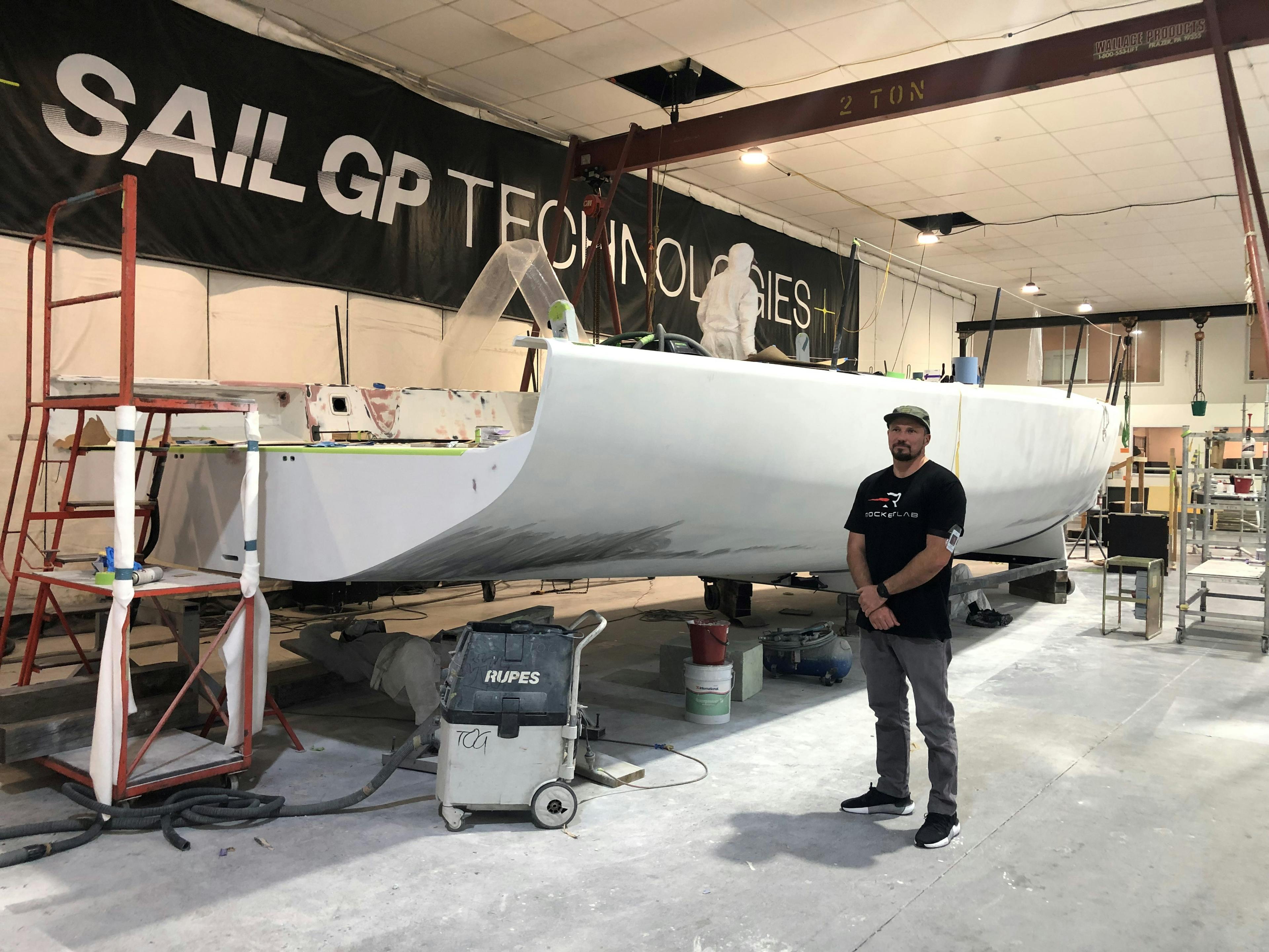 Malcolm stands next to a large, white, metal structure. It's held up on a platform and still under construction. In the background is a sign saying "SAILGP TECHNOLOGIES"