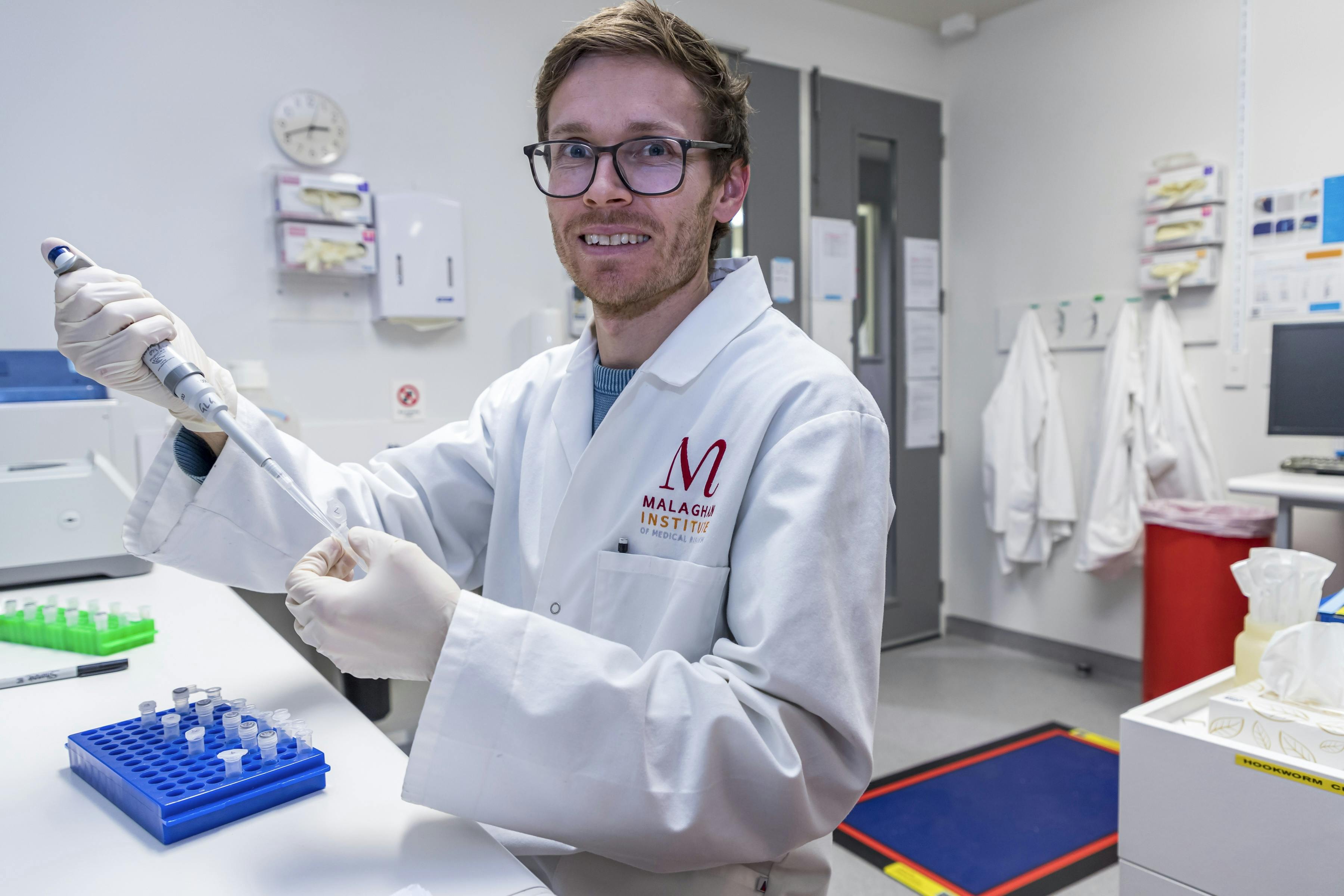 A man in a white lab coat and rubber gloves holds a pipette up to an eppendorf tube in a lab.