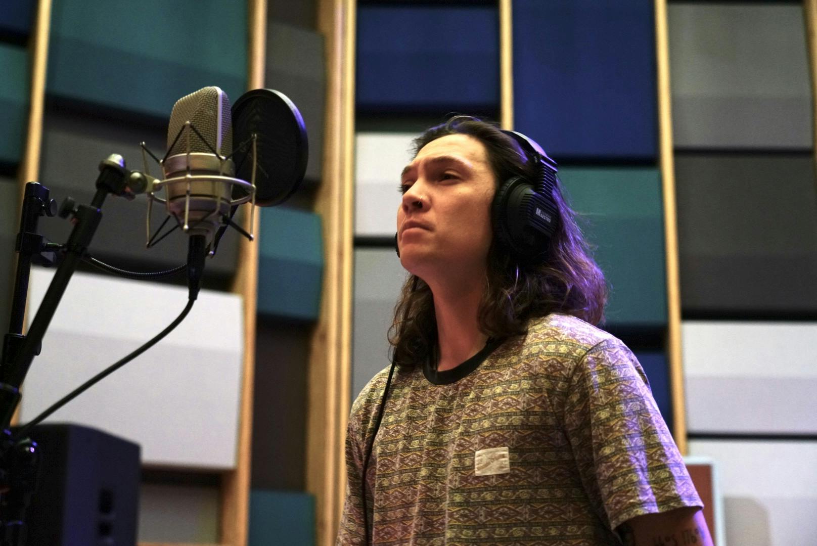 A man with long hair wearing a t-shirt and headphones stands in front of a microphone.